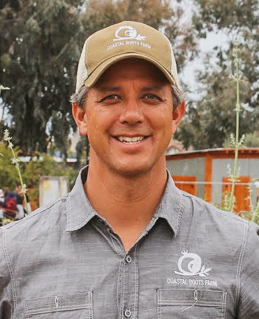 Smiling man wearing Coastal Roots shirt and hat with a farm in the background