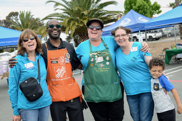 Four smiling adults and one child with their arms around each other. In the background are UC Master Gardener Program of San Diego tent canopies and palm trees.