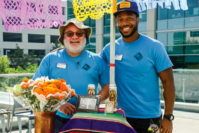 Two smiling UC Master Gardeners standing behind a circular ofrenda table brightly decorated with a Mexican blanket and orange flowers. In the background are San DIego County Operations Center buildings.