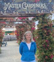 UC Master Gardener Jodi Bay standing in front of a Garden entrance with a large colorful sign saying Master Gardeners